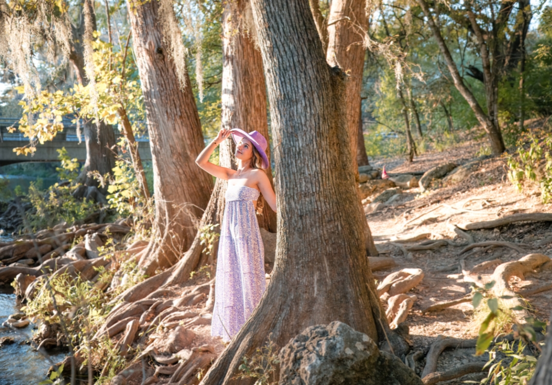 High school senior in purple and white dress with hat, posing by the Guadalupe River bank.