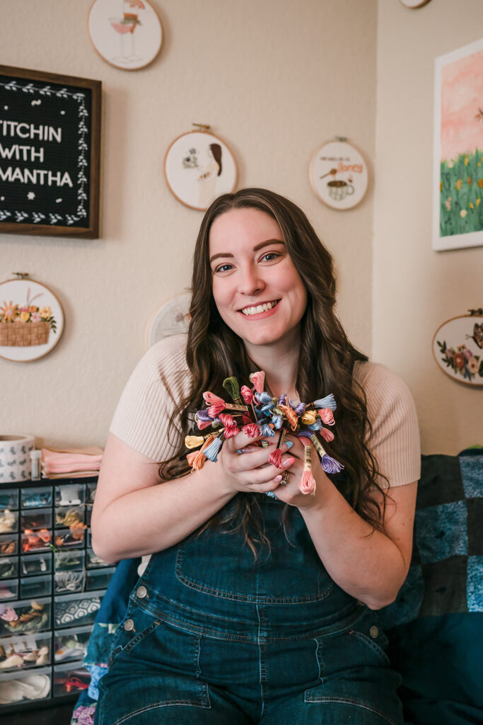 Woman smiling and holding embroidery thread bundles in her home studio.