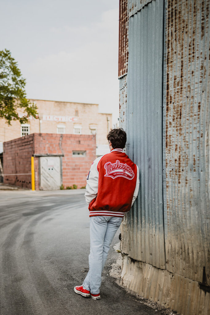 High School Senior leaning against building wearing letterman jacket